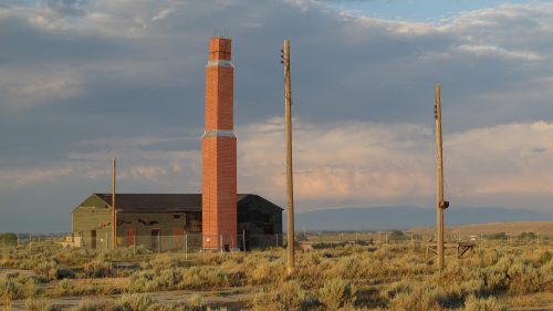 An old, weathered building with a tall chimney stands in a barren landscape under a cloudy sky.