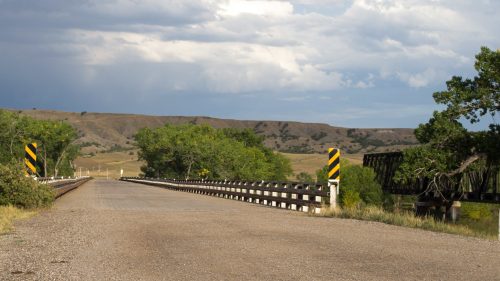 A gravel road leads to a wooden bridge, surrounded by green trees and rolling hills under a cloudy sky.