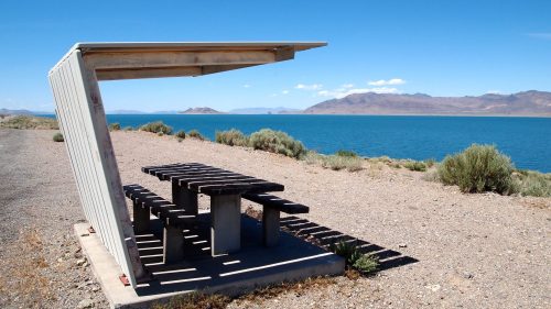 A shaded picnic table by a lake, surrounded by shrubs and mountains under a clear blue sky.