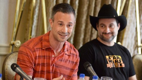 Two men sit at a table with microphones, one in a red striped shirt and the other in a black cowboy hat and t-shirt.