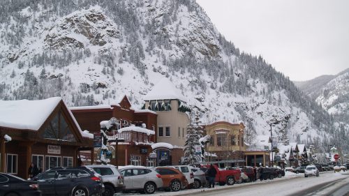 Snow-covered mountain backdrop with a quaint town street lined with shops and parked cars.