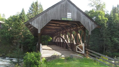 A wooden covered bridge named Bacon Bridge, surrounded by trees and a river, with a "Bridge Closed" sign.