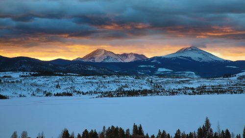 Snow-covered mountains under a dramatic sky at sunset, with a frozen lake in the foreground.