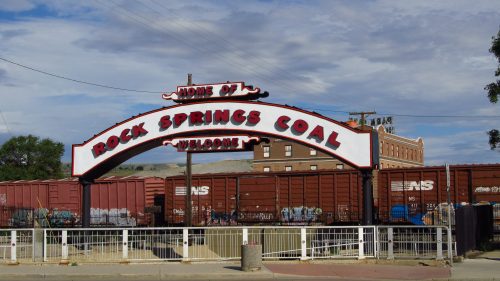 Sign reading "Home of Rock Springs Coal" with freight train cars in the background under a cloudy sky.