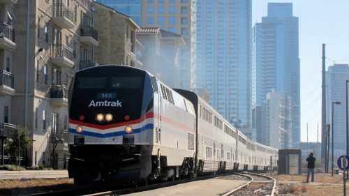An Amtrak train travels through a cityscape with tall buildings in the background on a clear day.