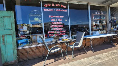 A storefront displaying antiques and collectibles, with tables and chairs outside under a bright blue sky.