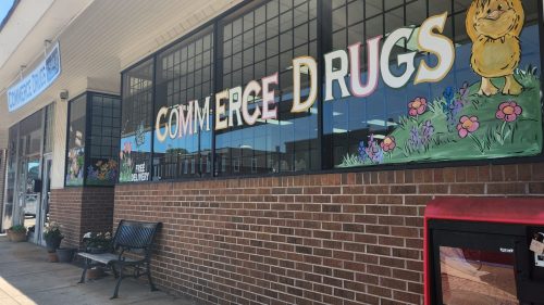 A storefront with "Commerce Drugs" painted on the windows, featuring flowers and a bench outside.