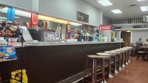 A cozy diner interior with a counter, stools, and a cashier serving customers in a casual setting.