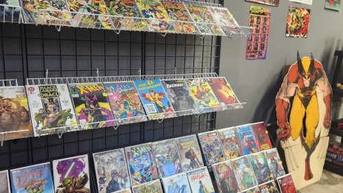 A display of comic books on shelves, featuring various titles and a large superhero cutout in the background.