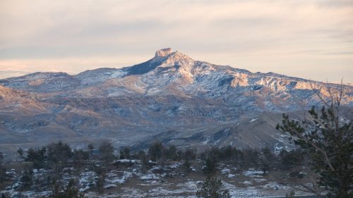 A snow-capped mountain rises above a landscape of rolling hills and sparse trees under a cloudy sky.