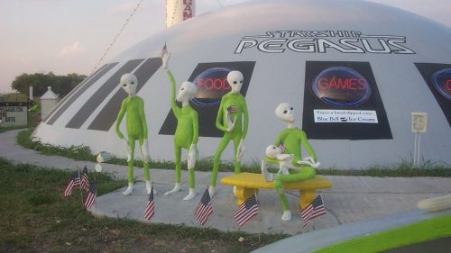 Four green aliens stand outside a dome-shaped building, holding small American flags, with a sign for food and games.