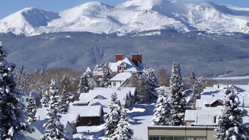 Snow-covered mountains loom over a quaint village with houses and trees blanketed in white under a clear blue sky.
