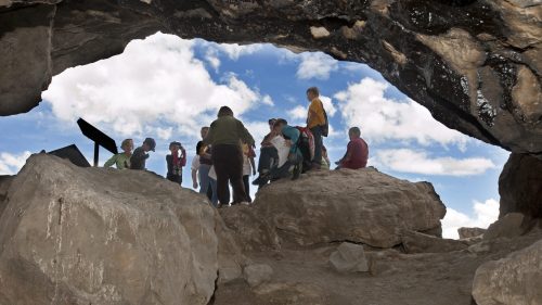 A group of people gathered inside a cave, with bright blue skies visible through the opening above.