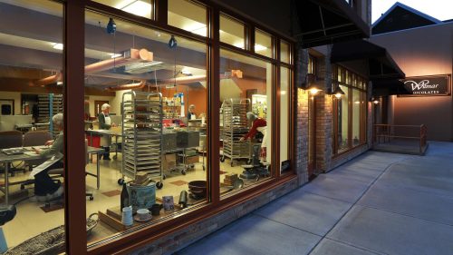 A bakery with large windows showing bakers at work, surrounded by baking trays and equipment. Evening light illuminates the scene.
