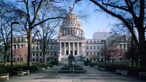 A grand government building with a dome, surrounded by trees and a statue in the foreground, under a clear blue sky.