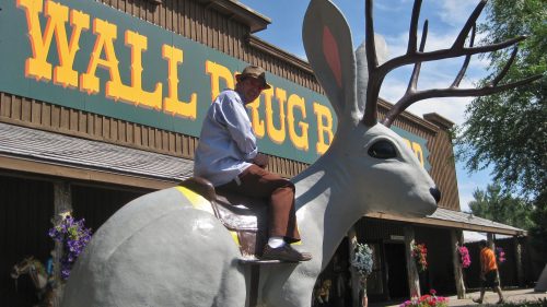 A man sits on a large, whimsical jackalope statue outside Wall Drug Store, with colorful flowers in the foreground.