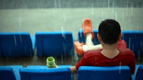 A person in a red shirt and orange clogs sits on blue seats, holding a cup, as rain falls around them.
