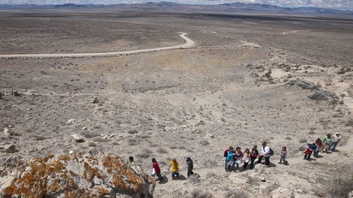 A group of people hiking down a rocky slope in a vast, arid landscape under a cloudy sky.