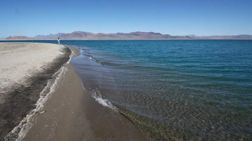 A serene beach with gentle waves lapping at the shore, leading to distant mountains under a clear blue sky.