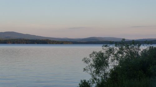 A serene lake surrounded by mountains, with calm water reflecting the sky and lush greenery in the foreground.