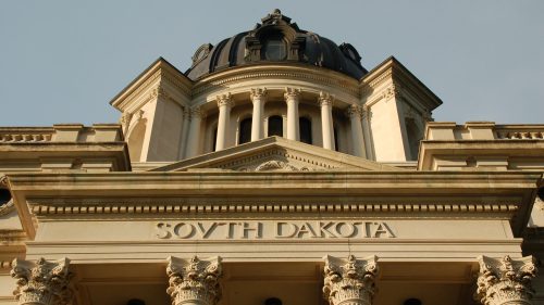 The ornate facade of the South Dakota State Capitol building, featuring columns and a dome against a clear sky.