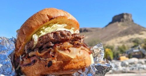 A piled high burger held up to the backdrop of Colorado from Bob's Atomic Burgers.