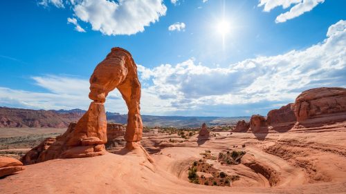 A stunning view of a natural rock arch under a bright blue sky with scattered clouds and sun shining in the background.