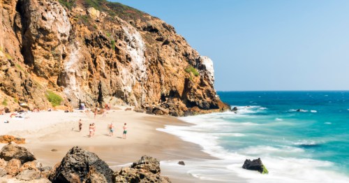 People walk toward the bright blue ocean and next to cliff walls on Zuma Beach in Southern California.