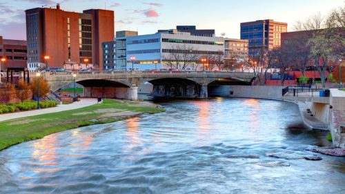 A river flows through a cityscape at sunset, with buildings and a bridge in the background.