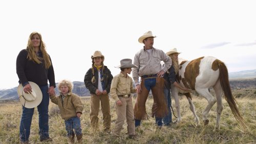 A family in cowboy attire stands in a grassy field with a horse, set against a cloudy sky and distant mountains.