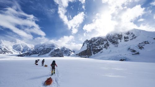 A group of climbers trekking across a snowy landscape with mountains and a bright sky in the background.