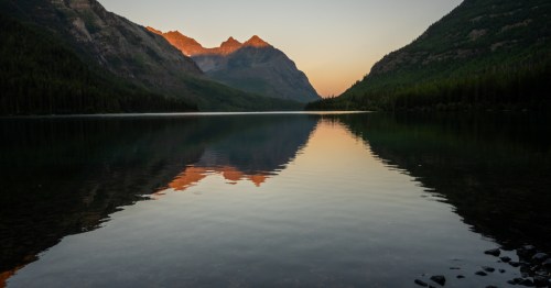 Mountains are reflected in the calm water of Kintla Lake at sunrise.