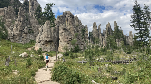 A hiker walks along a trail surrounded by towering rock formations and lush greenery under a cloudy sky.