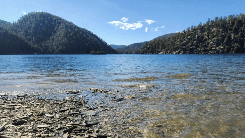 A serene lake surrounded by mountains, with clear water and rocky shoreline under a blue sky.