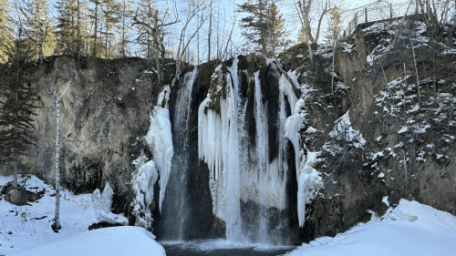A frozen waterfall surrounded by snow-covered rocks and trees, with icicles hanging from the cliff above.