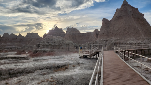 A wooden walkway leads through rugged, rocky formations under a cloudy sky at sunset in a desert landscape.