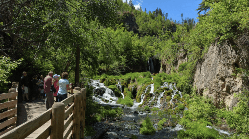 A scenic view of a waterfall surrounded by lush greenery, with people walking along a wooden path nearby.