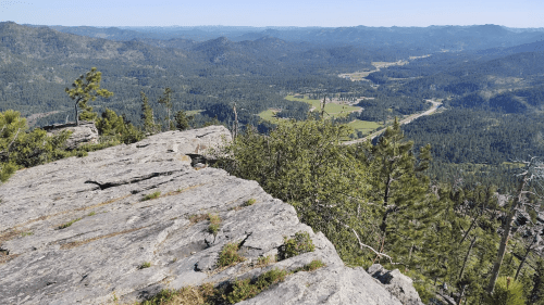 A panoramic view from a rocky outcrop, overlooking lush green valleys and distant mountains under a clear blue sky.