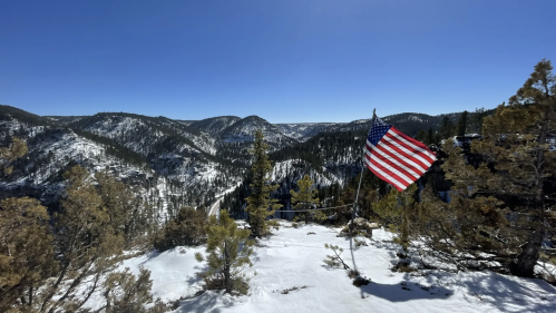 A snowy mountain landscape with a waving American flag in the foreground against a clear blue sky.