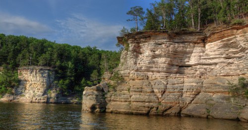 Sandstone cliffs stand on a river's edge topped with green trees.
