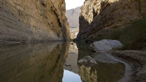 A serene river flows between towering rock formations, reflecting the cliffs in calm water.