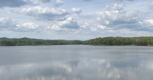Wide view of Deam Lake, Indiana reflecting white clouds and blue sky surrounded by green forest.