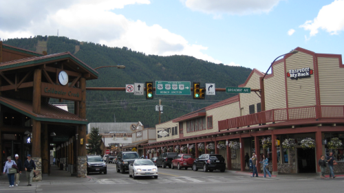 A street view of a small town with shops, traffic lights, and mountains in the background under a cloudy sky.
