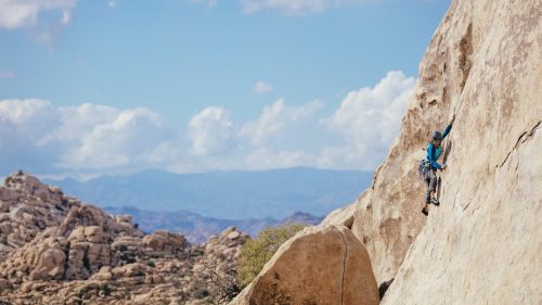 A climber ascends a rocky cliff in a desert landscape under a blue sky with fluffy clouds.