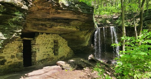 Historic stone moonshiner hideout beside a scenic waterfall on the Moonshiners Cave Trail in Arkansas surrounded by lush forest.