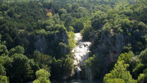 A scenic view of a waterfall cascading down rocky cliffs, surrounded by lush green trees and hills in the background.