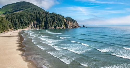 It's a sunny day at Short Sand Beach in Oregon. A long stretch of sand is seen with waves slowly rushing up towards it, and a mountain with trees sits in the background.