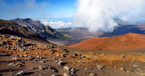 Maui's Haleakalā Volcano is seen from above, with clouds partially covering orange and red sands that border the Sliding Sands Trail.