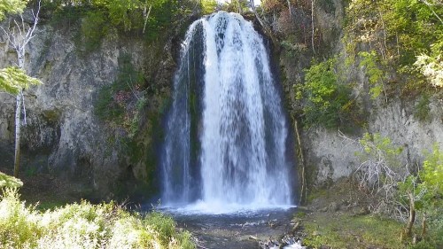A cascading waterfall surrounded by lush greenery and rocky cliffs, with sunlight filtering through the trees.