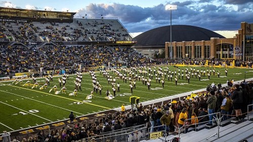 A large crowd watches a marching band perform on a football field at sunset, with stadium seating in the foreground.